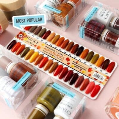 A selection of nail polish shades from the CHANCE Matching Trio - Hello Autumn Collection (36 Colors) is displayed on a swatch palette labeled "MOST POPULAR," surrounded by various bottles and packaging. The palette features autumn colors labeled "AUTUMN SPICE" and offers an opportunity to find your Matching Trio among 36 colors.
